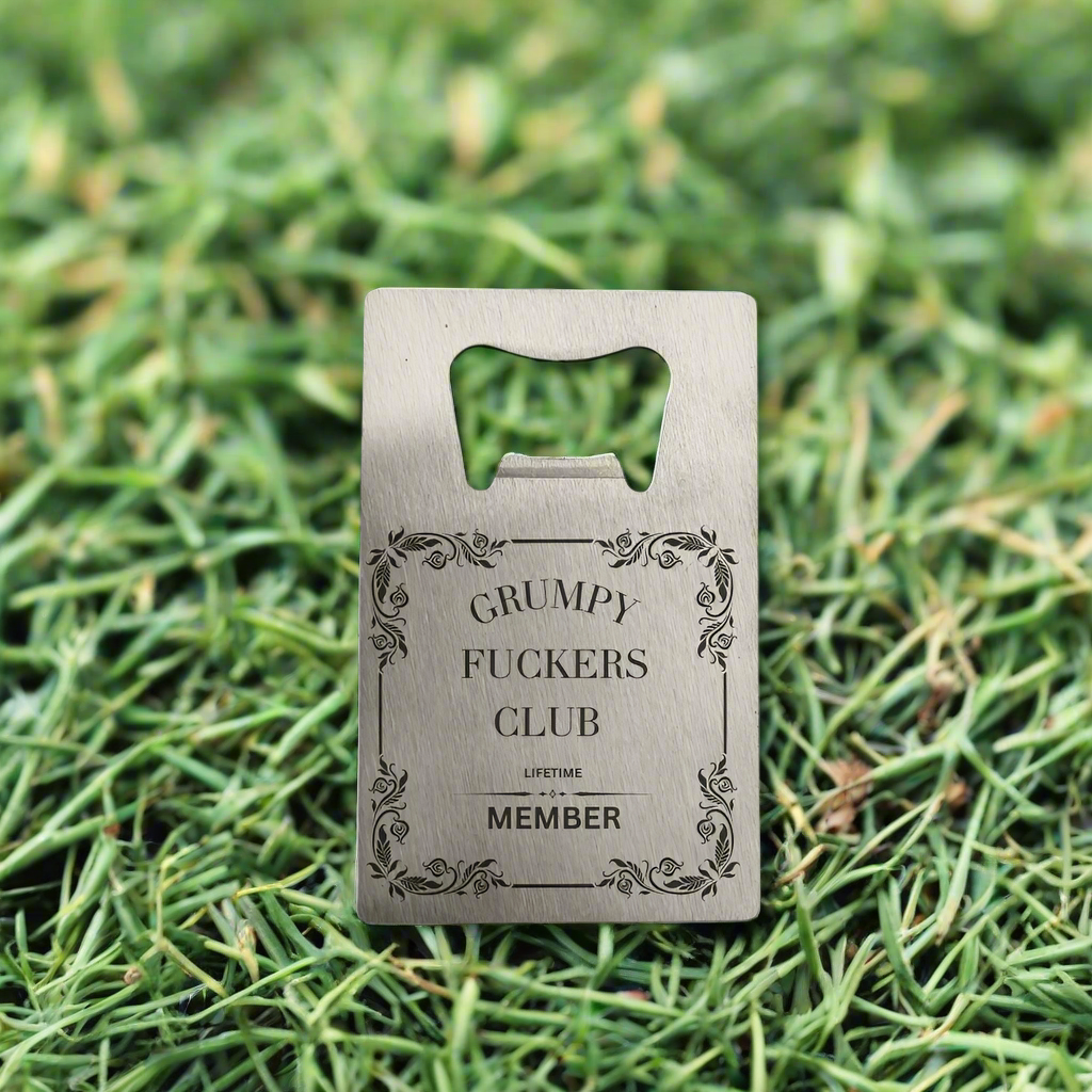 A stainless steel bottle opener with the text 'GRUMPY FUCKERS CLUB LIFETIME MEMBER' engraved on it, placed on a grassy background.