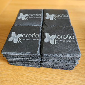 Four dark gray slate coasters stacked in two columns on a light wood surface. Each coaster features a white butterfly logo and the text 'icrotia K Proud to be me'.