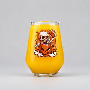 A clear stemless wine glass filled with orange juice, featuring a Halloween graphic of a skull inside a jack-o'-lantern mug, on a light gray background.