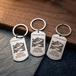 Three polished stainless steel dog tag keyrings engraved with "I Climbed Ben Nevis", a mountain illustration, the height 1345m and the date 26 May 2025, arranged on slate and walnut wood beside navy fabric
