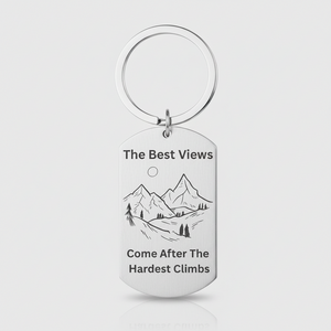 A silver metal dog tag keychain with a circular split ring, featuring a black engraved mountain illustration and the text 'The Best Views Come After The Hardest Climb' on a light gray background.