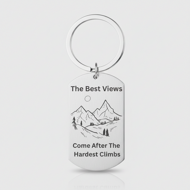 A silver metal dog tag keychain with a circular split ring, featuring a black engraved mountain illustration and the text 'The Best Views Come After The Hardest Climb' on a light gray background.
