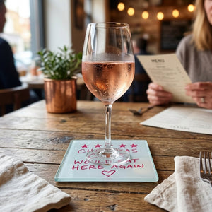 A square glass coaster with a white surface featuring red text that reads 'CHEF WAS CUTE, WOULD EAT HERE AGAIN' with five red stars at the top and a red heart below, shown on a table with a wine glass resting on top