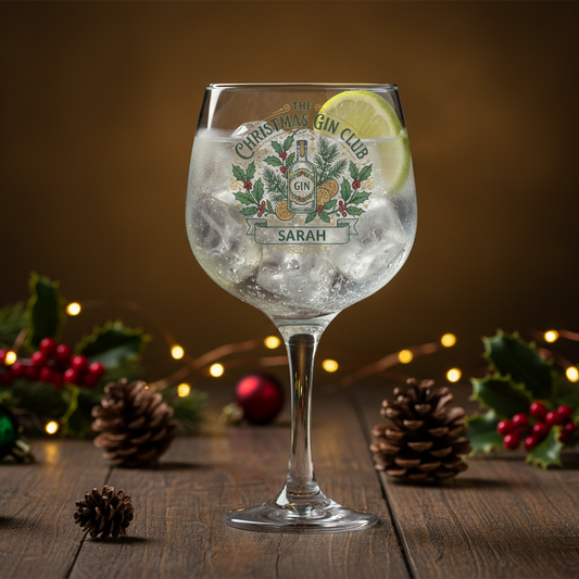 A clear stemmed glass with a festive Christmas-themed printed label reading 'The Christmas Gin Club' and the name 'SARAH', filled with a gin cocktail and ice, with a lemon slice garnish. The glass is set on a wooden table surrounded by pinecones, holly leaves, red berries, and warm string lights against a dark brown background.