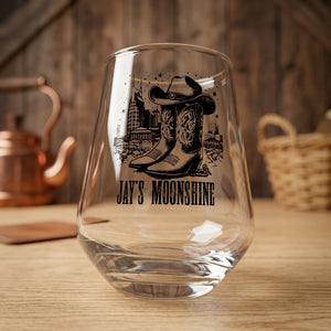 A clear stemless wine glass with a black western-themed graphic of cowboy boots and a hat, with the text 'JAY'S MOONSHINE' printed below, set on a wooden table with a rustic background.