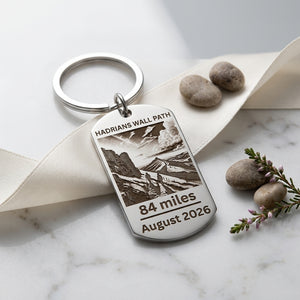 A personalised silver metal keychain with an engraved dog tag featuring a landscape illustration and the text 'HADRIANS WALL PATH', '84 miles', and 'August 2026'. The keychain is styled on a cream ribbon over a white marble surface with decorative stones and a sprig of purple flowers.