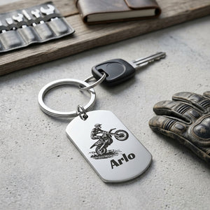 A silver metal keychain with a dog tag pendant engraved with a motorcycle rider silhouette and the name 'Arlo', shown with a key on a textured gray surface with motorcycle-themed props.