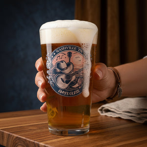 A hand holding a clear pint glass with a printed Nashville Tennessee theme graphic that reads 'Nashville TN' and 'Amy's Glass,' featuring a cowboy hat and guitar design, set on a wooden table.