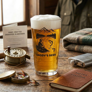 A personalised pint glass with a fishing design and personalised text daddys beer' on a table with fishing equipment surrounding 