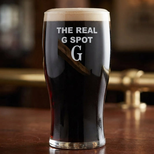 A dark beer glass with white text reading 'THE REAL G SPOT' and a large 'G' on the front, filled with black beer and topped with a foamy head, set on a wooden bar in a blurred pub environment.