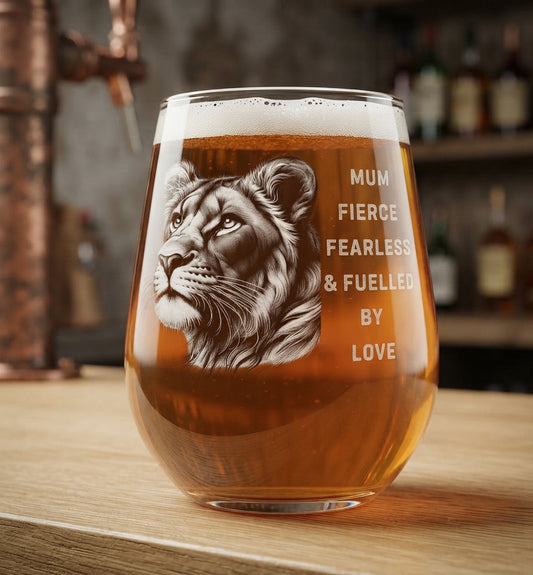A stemless wine glass filled with amber beer, featuring a detailed lion engraving and the text 'MUM FIERCE FEARLESS & FUELLED BY LOVE', set on a wooden bar counter with a blurred bar background.