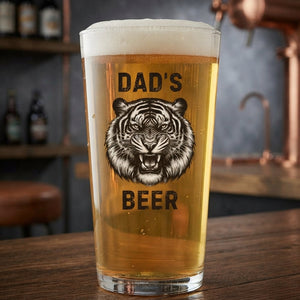 A pint glass filled with golden beer sits on a wooden bar counter. The glass features a black graphic of a tiger's face with the words 'DAD'S BEER' printed around it.