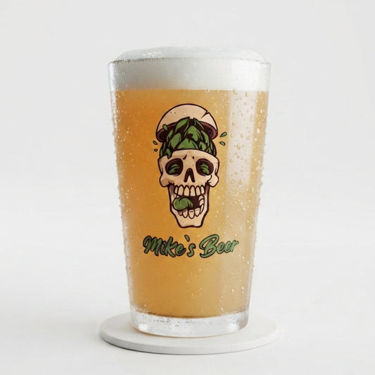 A clear pint glass filled with golden beer and topped with white foam, featuring a printed skull graphic with green pine branches and the text 'Miko's Bar' on the front, set on a white coaster against a light gray background.