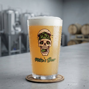 A frosted pint glass filled with golden beer and topped with white foam, standing on a wooden coaster on a gray bar counter. The glass features a cartoon skull graphic with a green beer hat and the text 'Mike's Bar'.