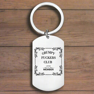 A silver keyring with the phrase 'GRUMPY FUCKERS CLUB LIFETIME MEMBER' printed in bold letters, set against a wooden background.
