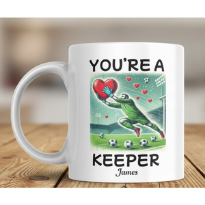 A white ceramic mug with a custom graphic print featuring a soccer goalkeeper diving to save a red heart, with the text 'YOU'RE A KEEPER James' on a wooden table with a blurred background.