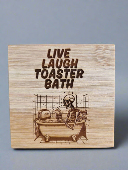 Laser engraved bamboo coaster with “Live Laugh Toaster Bath” text and skeleton in bathtub with toaster – dark humor eco-friendly gift, goth or emo room décor