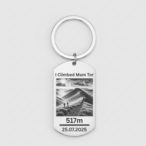 A silver metal dog tag keychain with a circular key ring, featuring a black and white photo of a mountain landscape with the text 'I Climbed Mam Tor', '517m', and '25.07.