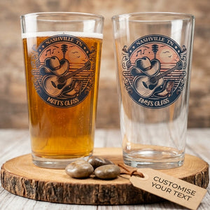 Two clear pint glasses displayed on a rustic wood slice, one filled with amber beer and one empty. Both glasses feature a circular Nashville Tix graphic with a guitar and the words 'Mats's Glass' and 'Pims Glass.