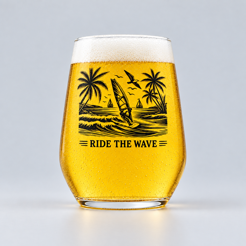 A clear stemless beer glass filled with golden beer and topped with white foam, featuring a black tropical wave graphic with palm trees, a windsurfer, birds, and the text 'RIDE THE WAVE' on the front, photographed on a light gray studio background.