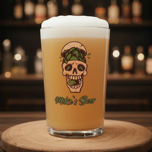 Personalised skull pint glass filled with beer, custom ink printed with personalised text in green script font, featuring a skull and hop crown illustration, displayed on a wooden board with a bar backdrop