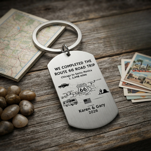 A personalized silver metal keychain shaped like a dog tag, engraved with a Route 66 road trip design and the names 'Karen & Gary 2026', displayed on a rustic wooden tabletop with a map, stones, and postcards nearby.