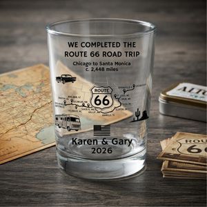 A clear glass shot tumbler with black printed Route 66 travel-themed text and illustrations, including a map, camper van, truck, cactus, and the names 'Karen & Gary' with '2026'. The glass is photographed on a dark wood surface with a vintage map, a metal tin, and a stack of Route 66 cards around it.