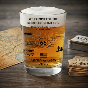A beer glass shot tumbler with black printed Route 66 travel-themed text and illustrations, including a map, camper van, truck, cactus, and the names 'Karen & Gary' with '2026'. The glass is photographed on a dark wood surface with a vintage map, a metal tin, and a stack of Route 66 cards around it.