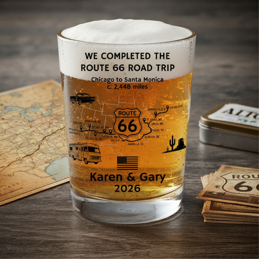 A beer glass shot tumbler with black printed Route 66 travel-themed text and illustrations, including a map, camper van, truck, cactus, and the names 'Karen & Gary' with '2026'. The glass is photographed on a dark wood surface with a vintage map, a metal tin, and a stack of Route 66 cards around it.