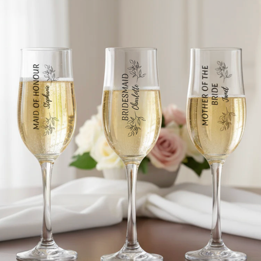 Three personalised stemmed champagne flutes filled with champagne, each printed with a different bridal party role and name — Maid of Honour, Bridesmaid and Mother of the Bride — styled with white roses and soft linen background