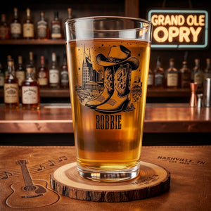 A clear pint glass filled with amber beer, featuring a black cowboy boots and hat graphic with the name 'ROBBIE' printed below, set on a wooden bar coaster in a bar environment with bottles and a neon sign in the background.