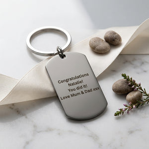A silver metal dog tag keychain with engraved personalised congratulations text, displayed on a cream ribbon over a white marble surface with decorative stones and a sprig of purple flowers.
