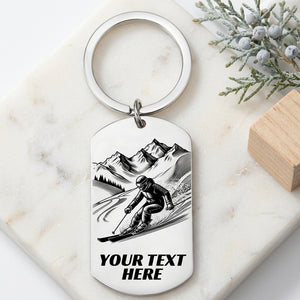A silver metal dog tag keychain with a ski graphic and the text 'YOUR TEXT HERE', styled on a white marble surface with decorative pine branches and a wooden block.
