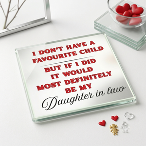 A square glass coaster with a humorous quote printed on it, styled on a white surface with small red heart decorations and a glass bowl in the background.
