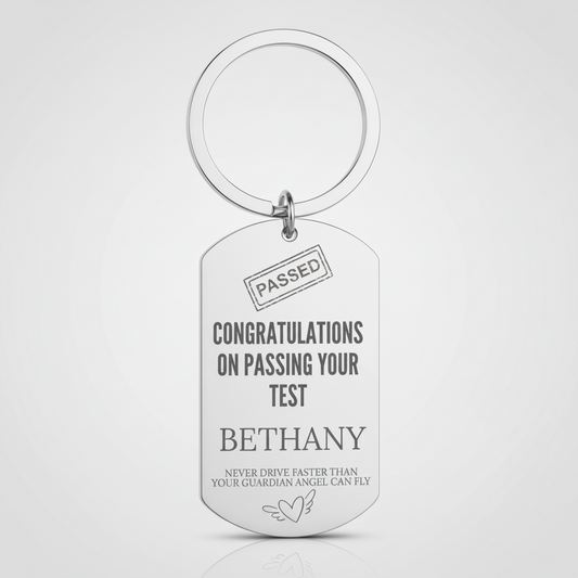 A silver metal dog tag keychain with a split key ring, featuring engraved personalized text that reads 'CONGRATULATIONS ON PASSING YOUR TEST BETHANY' and 'NEVER DRIVE FASTER THAN YOUR GUARDIAN ANGEL CAN FLY' on a light gray background.