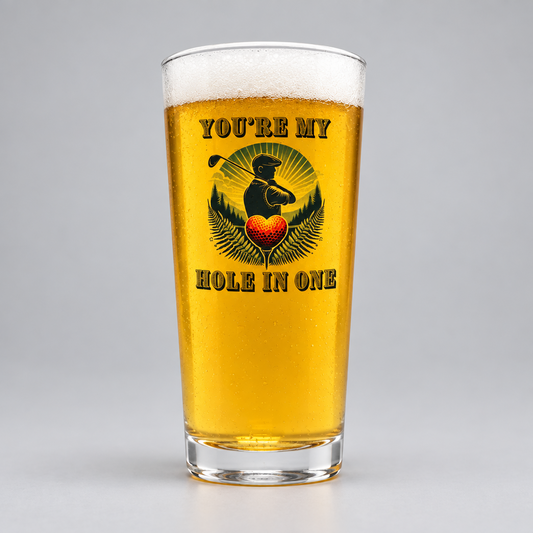 A clear pint glass filled with golden beer, featuring a printed graphic of a golfer silhouette, a red heart, and the text 'YOU'RE MY HOLE IN ONE' on a light gray studio background.
