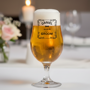Personalised stemmed tulip beer glass printed with name Daniel, wedding date and Groom role in ornate frame design, filled with beer and styled on a wedding table with roses and candle