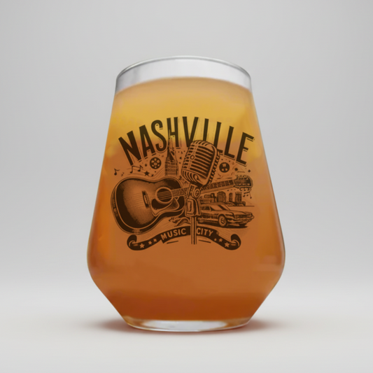 A stemless wine glass filled with an amber-colored beverage, featuring a black graphic print of a microphone, guitar, car, and the words 'NASHVILLE MUSIC CITY' on the front, set against a neutral gray background.