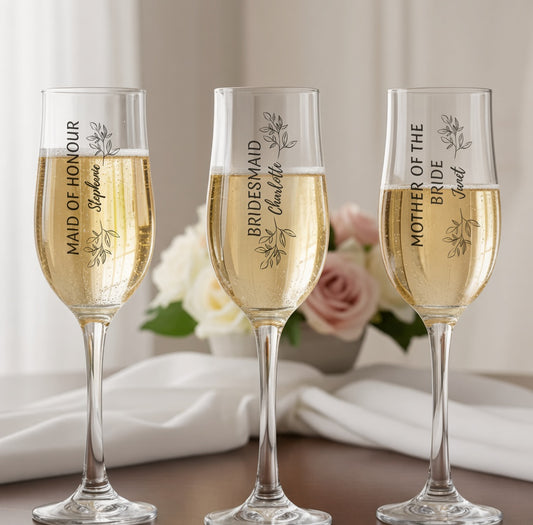 Three personalised stemmed champagne flutes filled with champagne, each printed with a different bridal party role and name — Maid of Honour, Bridesmaid and Mother of the Bride — styled with white roses and soft linen background