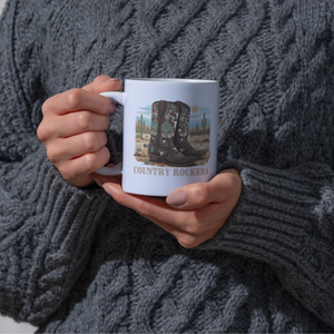 Person holding personalized cowboy boots mug while wearing gray knit 
sweater, showing custom text and desert landscape design on ceramic 
coffee cup, demonstrating daily use and personalization