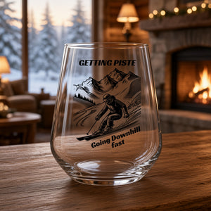A clear stemless wine glass with a black ski graphic and the text 'GETTING PISTE Going Downhill fast', displayed on a wooden table in a cozy, blurred living room with a fireplace and window view.