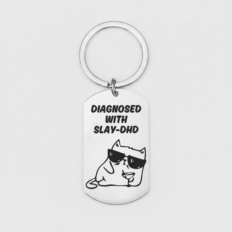 A silver metal dog tag keychain with a circular key ring, featuring a black graphic of a cartoon cat wearing sunglasses and the text 'DIAGNOSED WITH SLAY-DHD' on a light gray background.