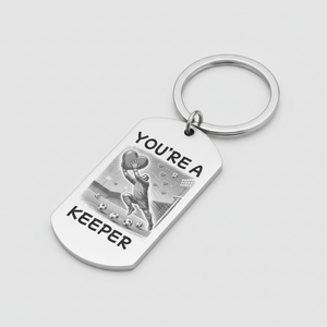 A silver metal dog tag keychain with a key ring, featuring a black and white illustration of a soccer player and the text 'YOU'RE A KEEPER' on a light gray background.